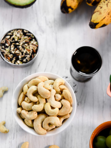 A little bowl of cashews with a few spilled on the table along with a bowl of olives, a measuring cup of wild rice, a little cup of olive oil and a bunch of bananas.