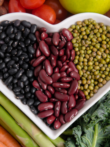 A heart shaped bowl filled with black beans, kidney beans and mung beans, surrounded by a variety of vegetables.