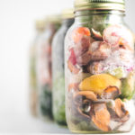 Four large mason jars full of frozen vegetable scraps for making vegetable broth, sitting in a diagonal line with the front jar in focus.