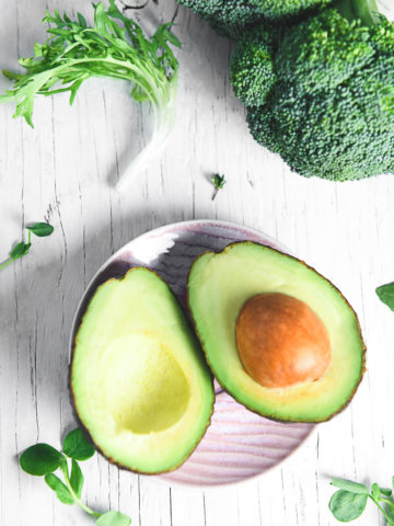 A sliced avocado lying open on a little pink plate surrounded by a variety of leafy greens, herbs and broccoli.