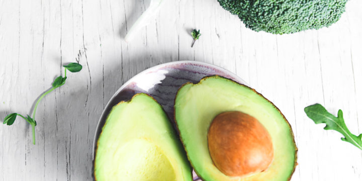 A sliced avocado lying open on a little pink plate surrounded by a variety of leafy greens, herbs and broccoli.