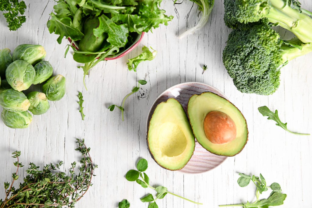 A sliced avocado lying open on a little pink plate surrounded by a variety of leafy greens, herbs and broccoli.