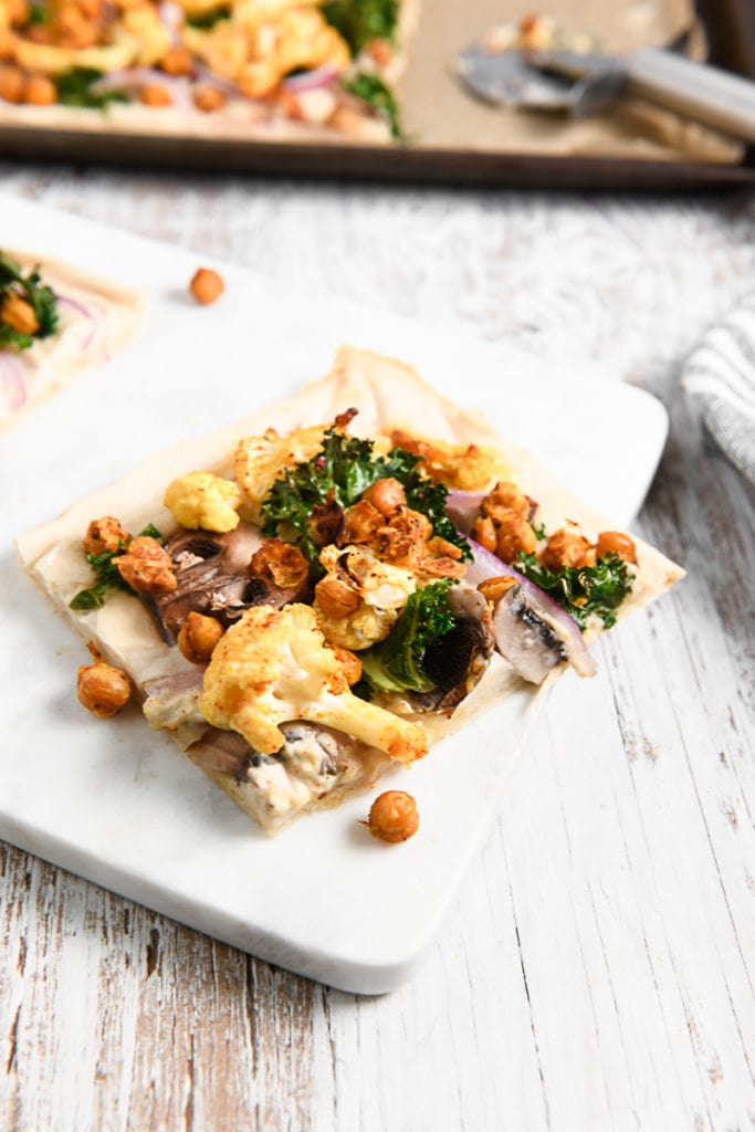 A slice of loaded buffalo chickpea flatbread pizza sitting on a cutting board in front of a baking sheet holding the rest of the pizza.