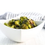 A bowl of roasted broccoli in a white square bowl with a striped linen cloth behind.