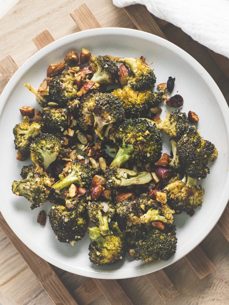 A plate of roasted broccoli, almonds and pumpkin seeds beside a white linen cloth.