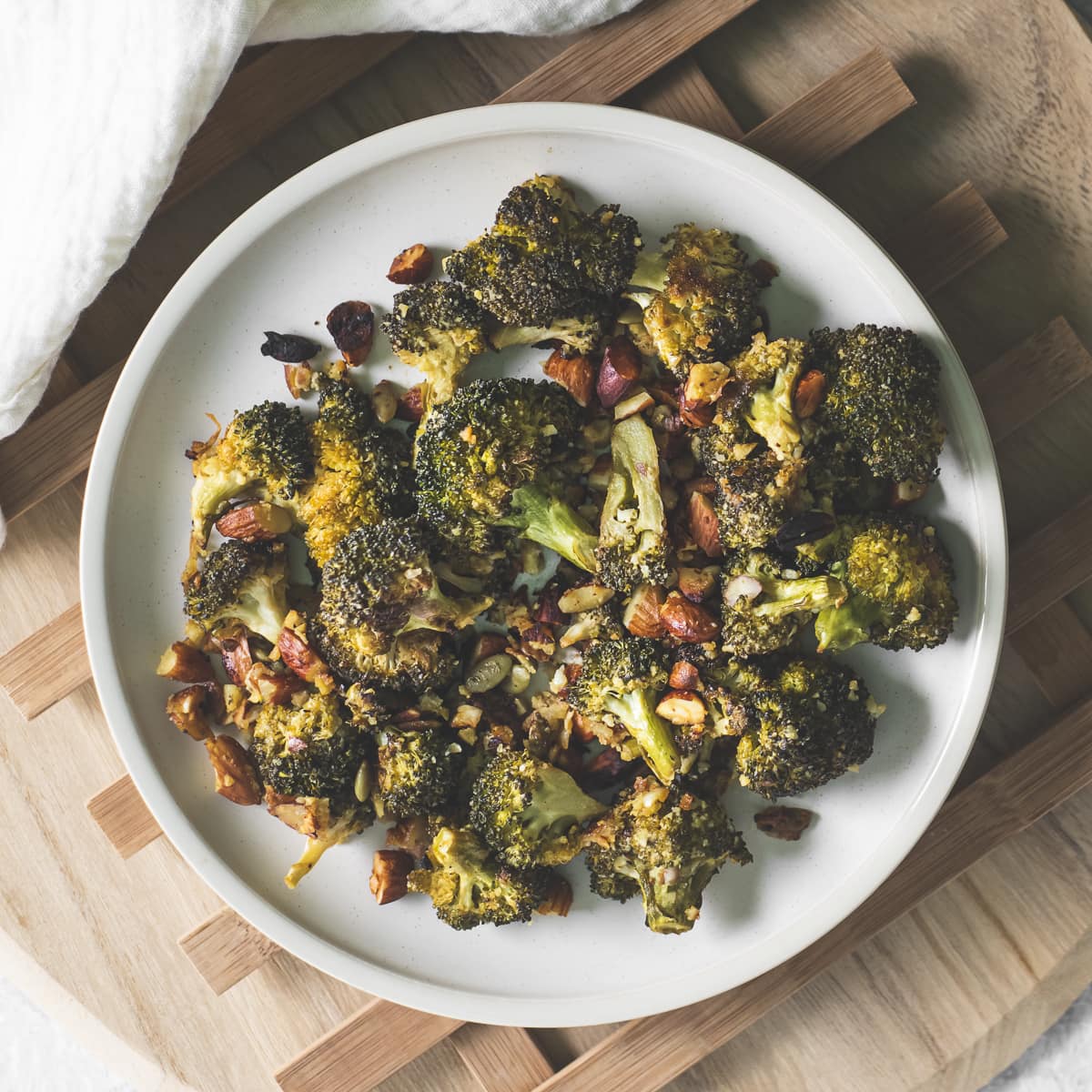 A plate of roasted broccoli, almonds and pumpkin seeds beside a white linen cloth.