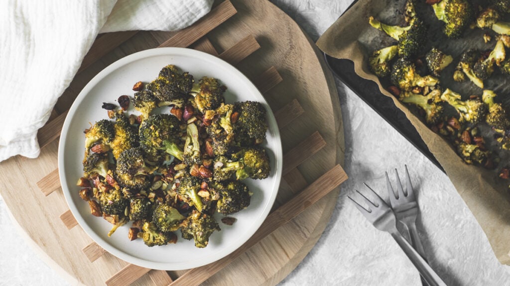 A plate of roasted broccoli and crispy chopped almonds beside a baking sheet, two forks and a linen napkin.