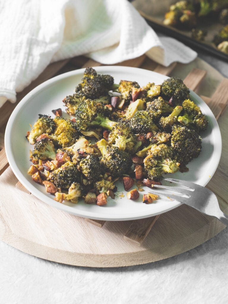 A plate of roasted broccoli and almonds with a fork resting on the side, a baking sheet in the background and linen napkin beside.