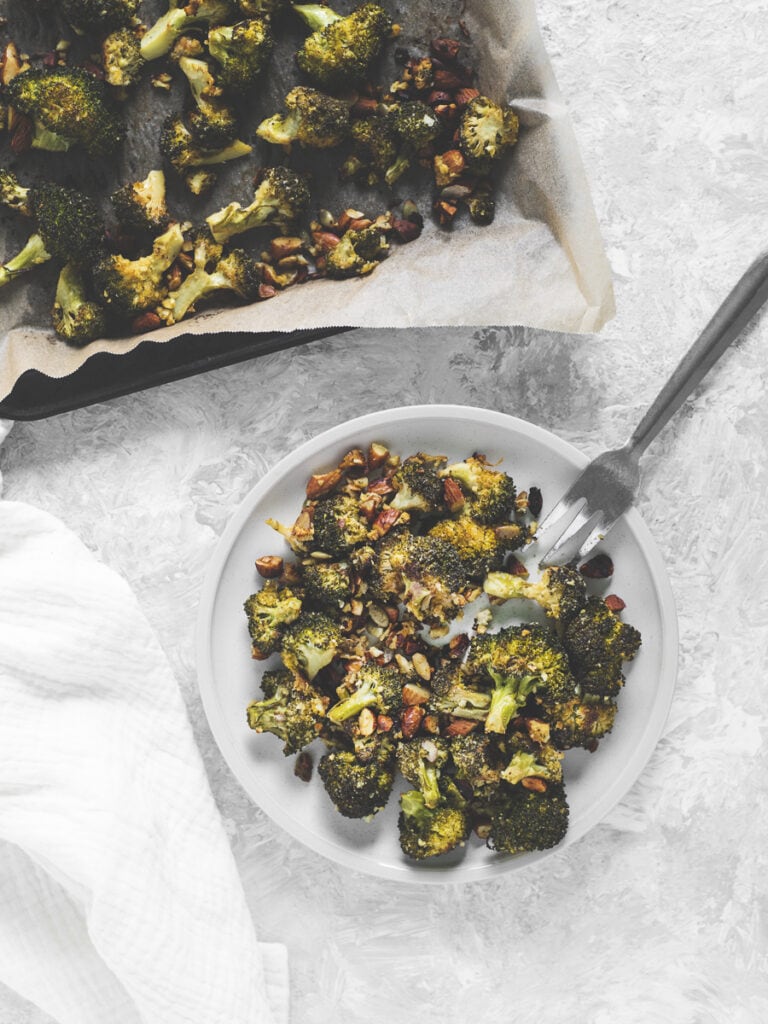 A plate of roasted broccoli, almonds and pumpkin seeds beside a full baking sheet and a linen cloth.