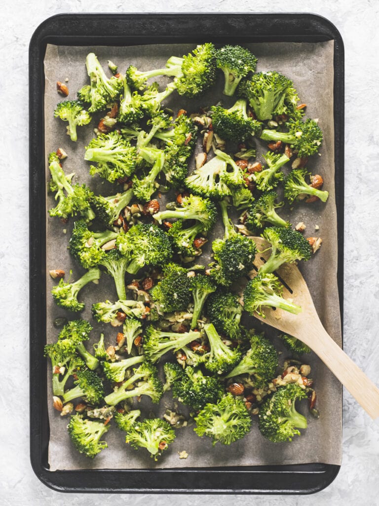 Broccoli florets and chopped almonds evenly spread on a baking sheet with a wooden spoon resting on the side.