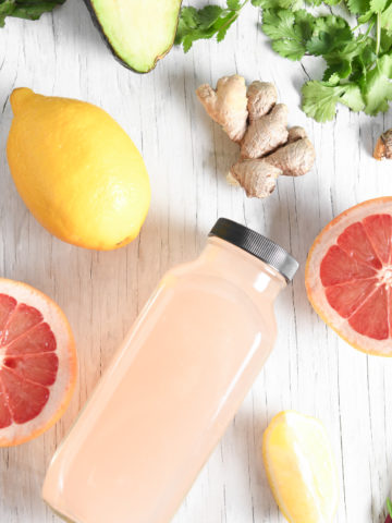 A bottle of pink grapefruit juice surrounded but a sliced open grapefruit, lemons, beets, greens and herbs.