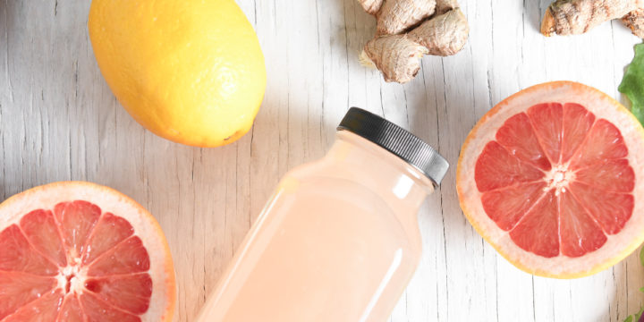 A bottle of pink grapefruit juice surrounded but a sliced open grapefruit, lemons, beets, greens and herbs.