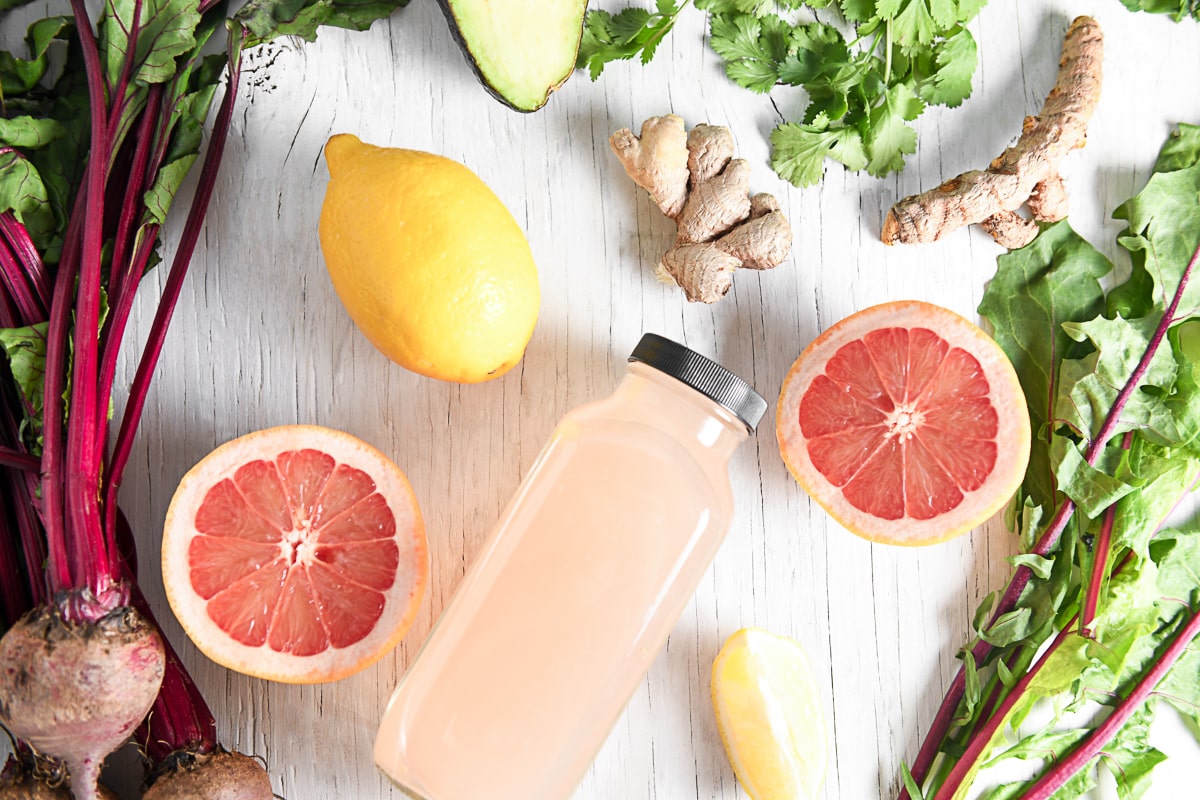 A bottle of pink grapefruit juice surrounded but a sliced open grapefruit, lemons, beets, greens and herbs.