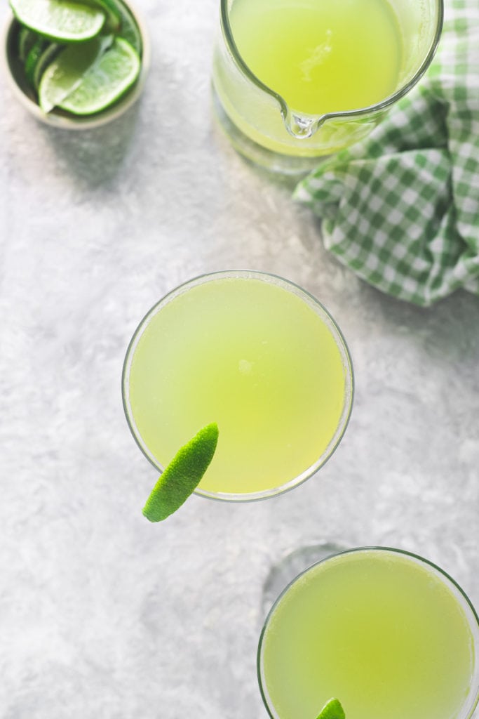 Two glasses filled with Green Apple Soda, garnished with limes, sitting beside a half empty pitcher and a bowl of lime wedges.