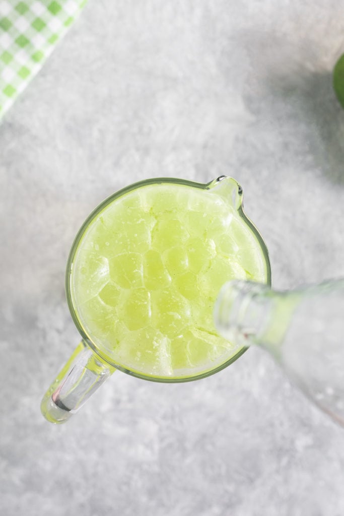 A bottle being emptied above a pitcher full of bubbling and foaming Green Apple and Lime Soda.