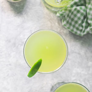 Two glasses filled with Green Apple Soda, garnished with limes, sitting beside a half empty pitcher and a bowl of lime wedges.