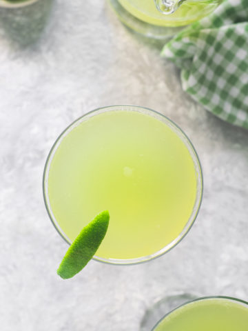 Two glasses filled with Green Apple Soda, garnished with limes, sitting beside a half empty pitcher and a bowl of lime wedges.