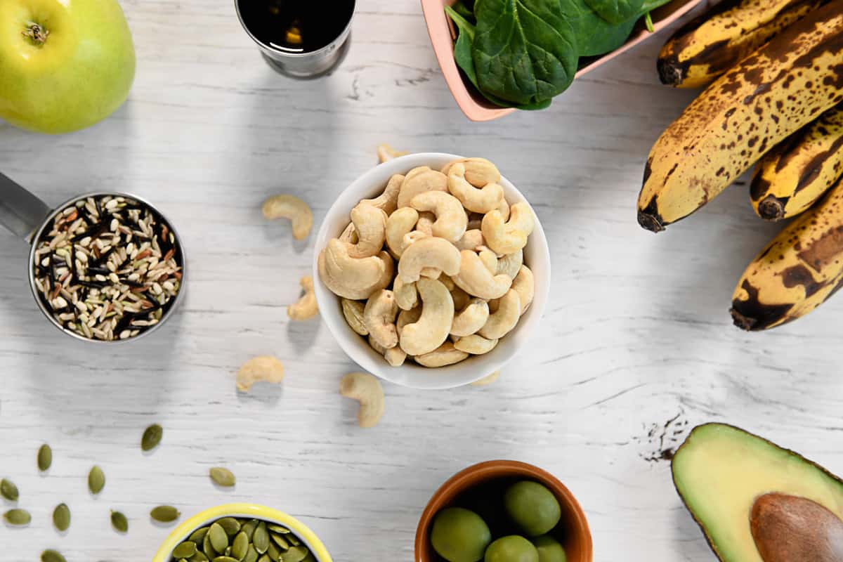 Whole foods including raw cashews, wild rice, olives, greens, fruits and a sliced avocado spread out on a rustic table.