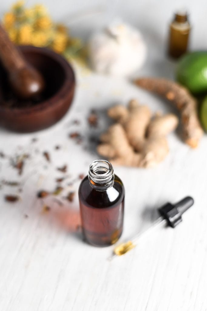 An open tincture bottle sitting in front of herbs, citrus fruits, ginger and a mortar and pestle.