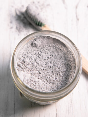 A quarter pint jar filled with charcoal toothpowder and a toothbrush that just been dipped in resting beside.