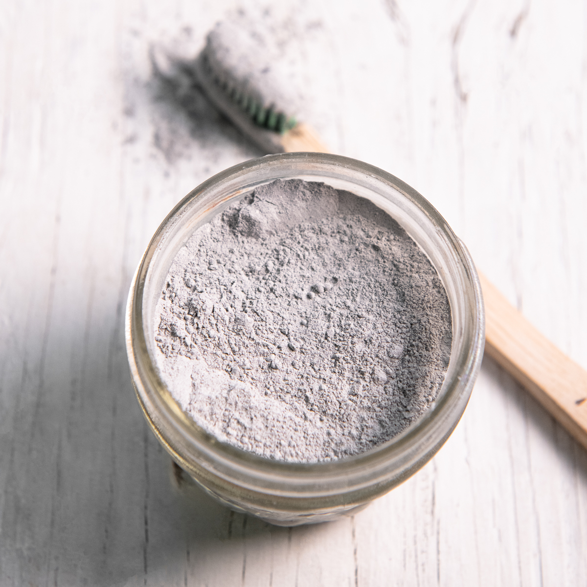 A quarter pint jar filled with charcoal toothpowder and a toothbrush that just been dipped in resting beside.