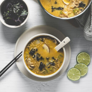 A freshly poured bowl of vegetable-filled Thai Noodle Soup beside a soup pot, bowl of kale and fresh limes.