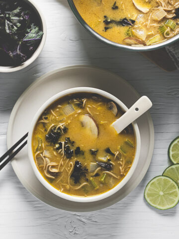 A freshly poured bowl of vegetable-filled Thai Noodle Soup beside a soup pot, bowl of kale and fresh limes.