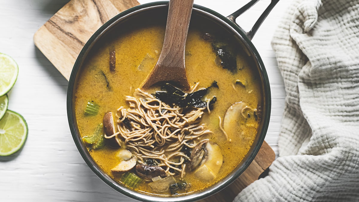 A wooden spoon stirring up a pot of Thai noodle soup with mushrooms, celery and kale.