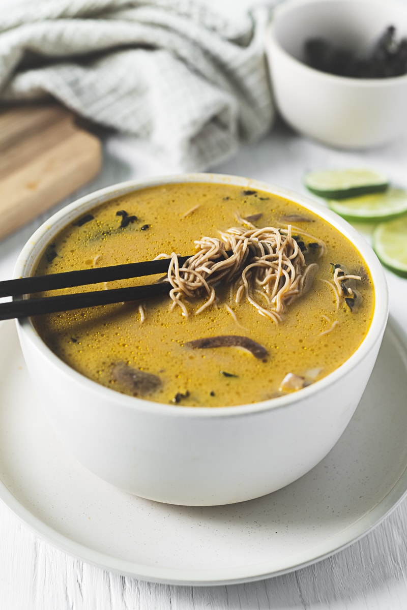 A large bowl of brothy coconut soup and a pair of chopsticks pulling up brown rice Thai noodles.