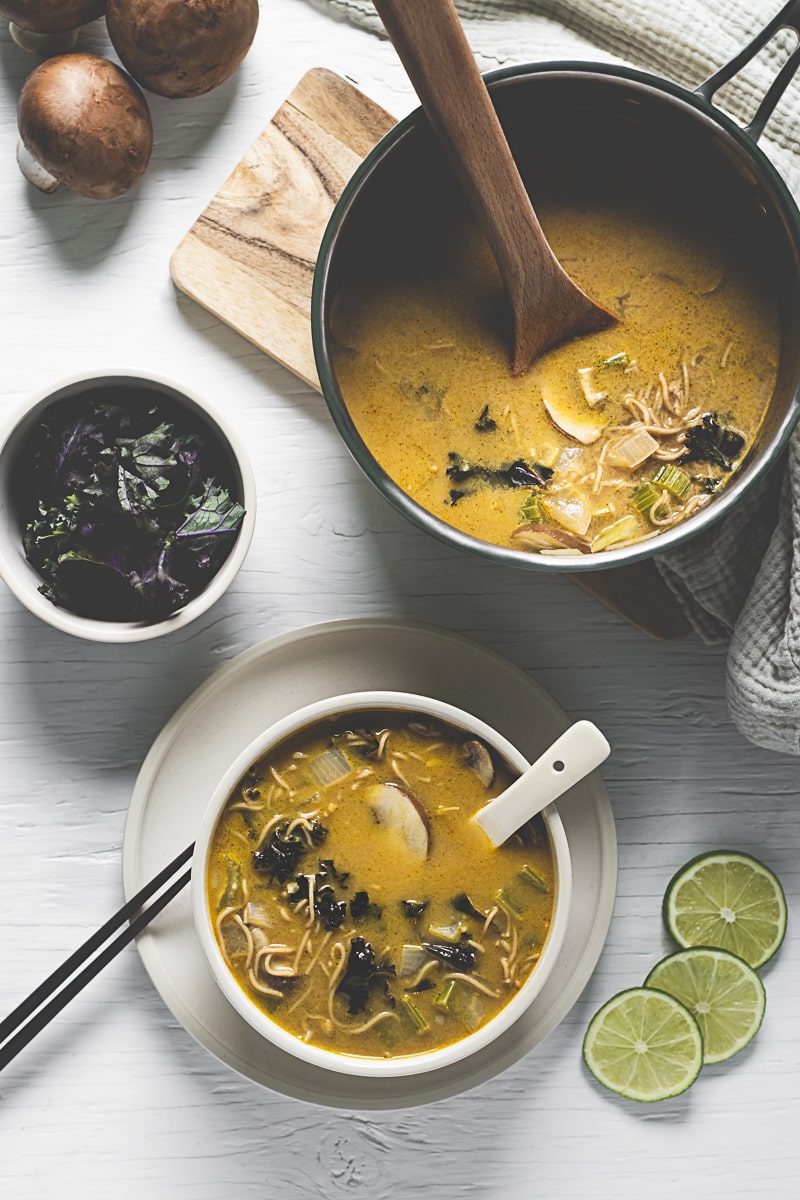 A freshly poured bowl of vegetable-filled Thai Noodle Soup beside a soup pot, bowl of kale and fresh limes. 