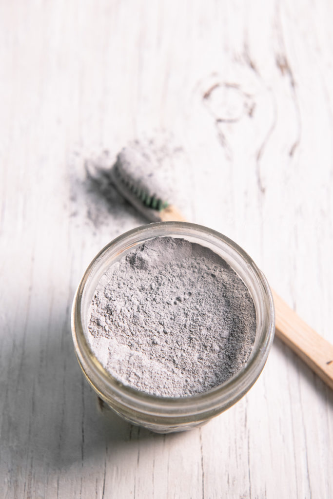 A quarter pint jar filled with charcoal toothpowder and a toothbrush that just been dipped in resting beside.