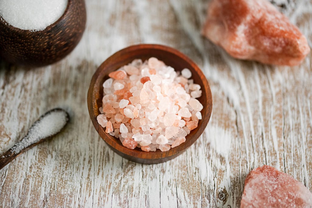 A heaping bowl of pink Himalayan salt surrounded by white table sat and rock salt.