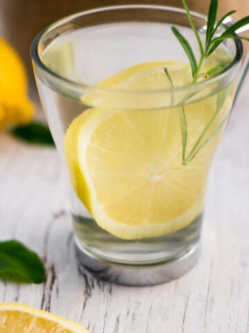 A clear mug with water, lemon and a sprig of rosemary,