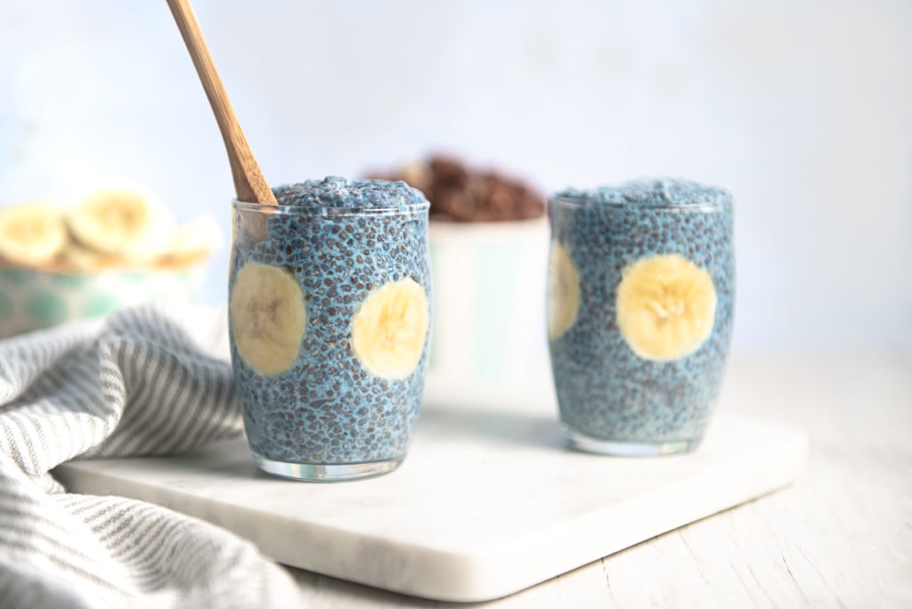 Two glass pudding cups full of blue chia pudding with bananas.