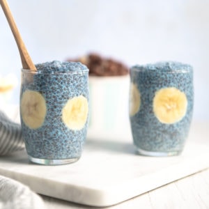 Two glass pudding cups full of blue chia pudding with bananas.