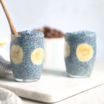 Two small glass pudding cups filled with Blue Majik chia pudding and banana slices, sitting on a marble cutting board with a white and grey linen napkin to the left and a bowl of nuts in the background.