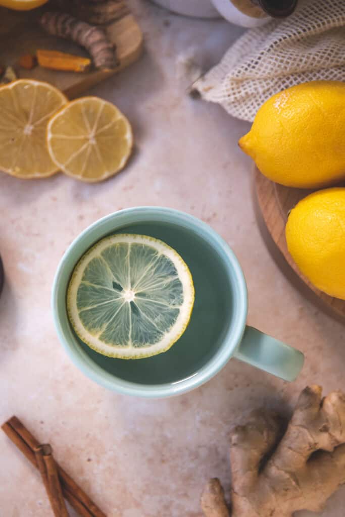 A lemon round floating in a teal mug surrounded by whole and sliced lemon, cinnamon sticks and ginger root.