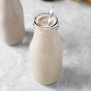 A cream coloured smoothie in a milk bottle with the tip of a white and grey straw peaking above the rim.