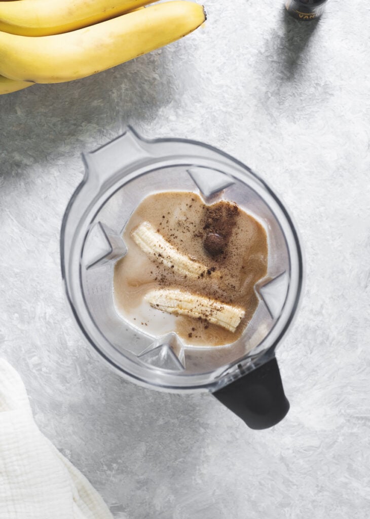 A blender jug filled with a ripe banana, milk and cinnamon spices, sitting in the middle of a linen cloth, a bunch of bananas and a bottle of vanilla extract.