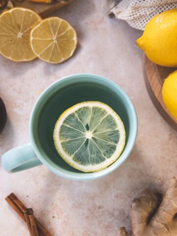 A sliced lemon round floating face-up in a teal mug surrounded by whole and sliced lemon, cinnamon sticks, ginger root and a honey dipper resting in a small bowl.