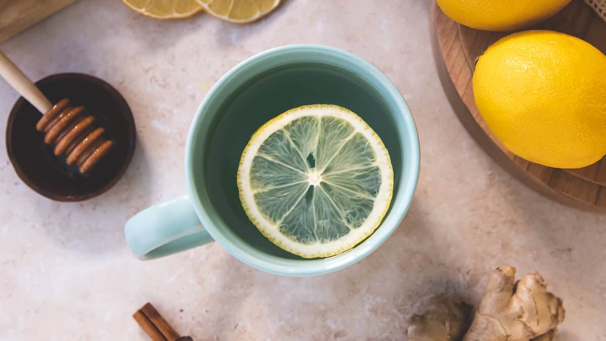 A sliced lemon round floating face-up in a teal mug surrounded by whole and sliced lemon, cinnamon sticks, ginger root and a honey dipper resting in a small bowl.