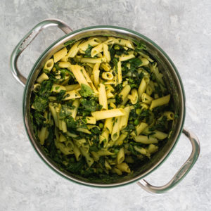 A large saucepan filled with cooked penne pasta tossed with spinach, peas, nutritional yeast, turmeric apple cider vinegar and healthy avocado oil.