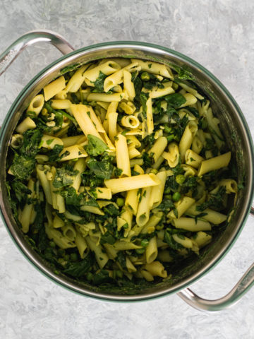 A large saucepan filled with cooked penne pasta tossed with spinach, peas, nutritional yeast, turmeric apple cider vinegar and healthy avocado oil.