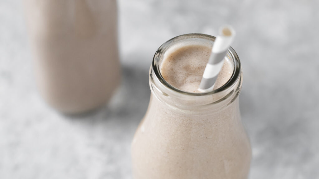 A cream coloured smoothie in a milk bottle with the tip of a white and grey straw peaking above the rim.