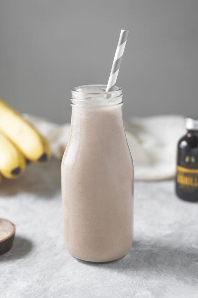 A creamy, vanilla smoothie in a single-serve milk jar with a striped straw sticking out to the right, sitting front and centre of the ingredients and linen cloth.