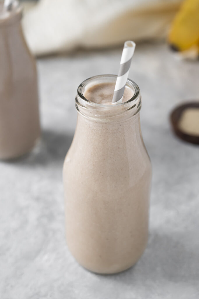 A jar filled with a creamy vanilla smoothie with a straw placed in, sitting in front of another smoothie in a jar, a bowl of ashwagandha powder and a bunch of bananas.