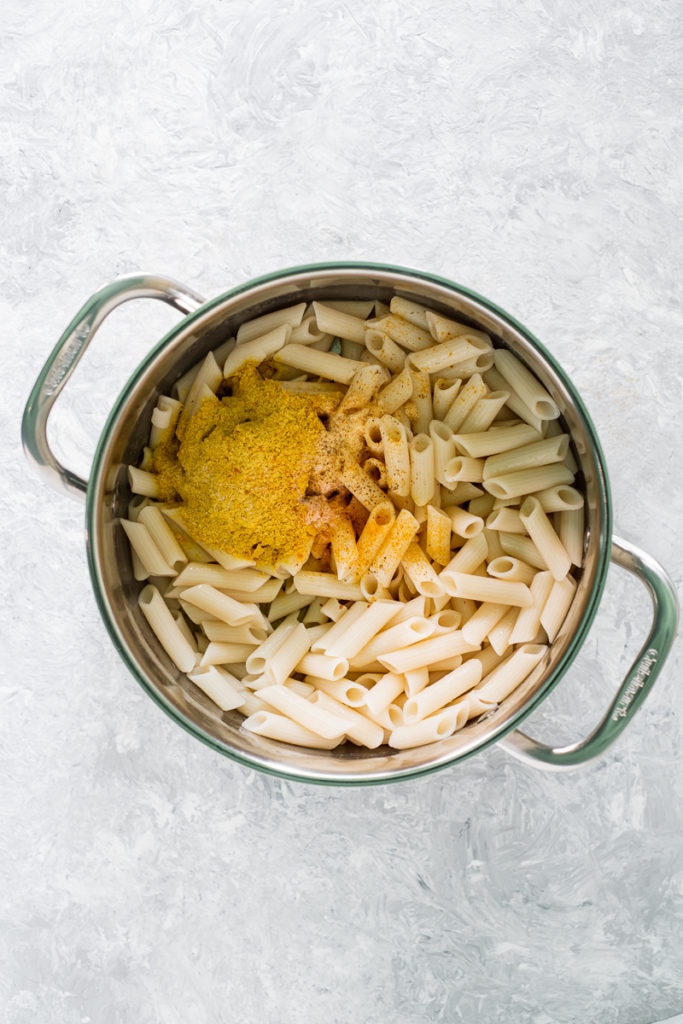 A large saucepan full of penne pasta with nutritional yeast, salt and turmeric poured on top.