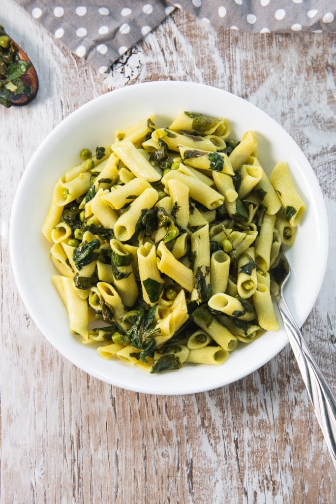 A heaping bowl of delicious Spinach and Peas Penne Pasta with a fork in the bowl and a polka dot napkin and wooden spoon with wilted spinach stuck to it sitting above.