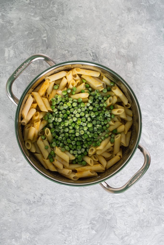 A large saucepan filled with penne pasta mixed with nutritional yeast, turmeric and apple cider vinegar with a cup of peas poured on top.