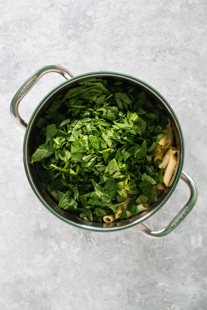 A large saucepan filled with penne pasta mixed with nutritional yeast, turmeric and apple cider vinegar with a heap of chopped spinach on top.